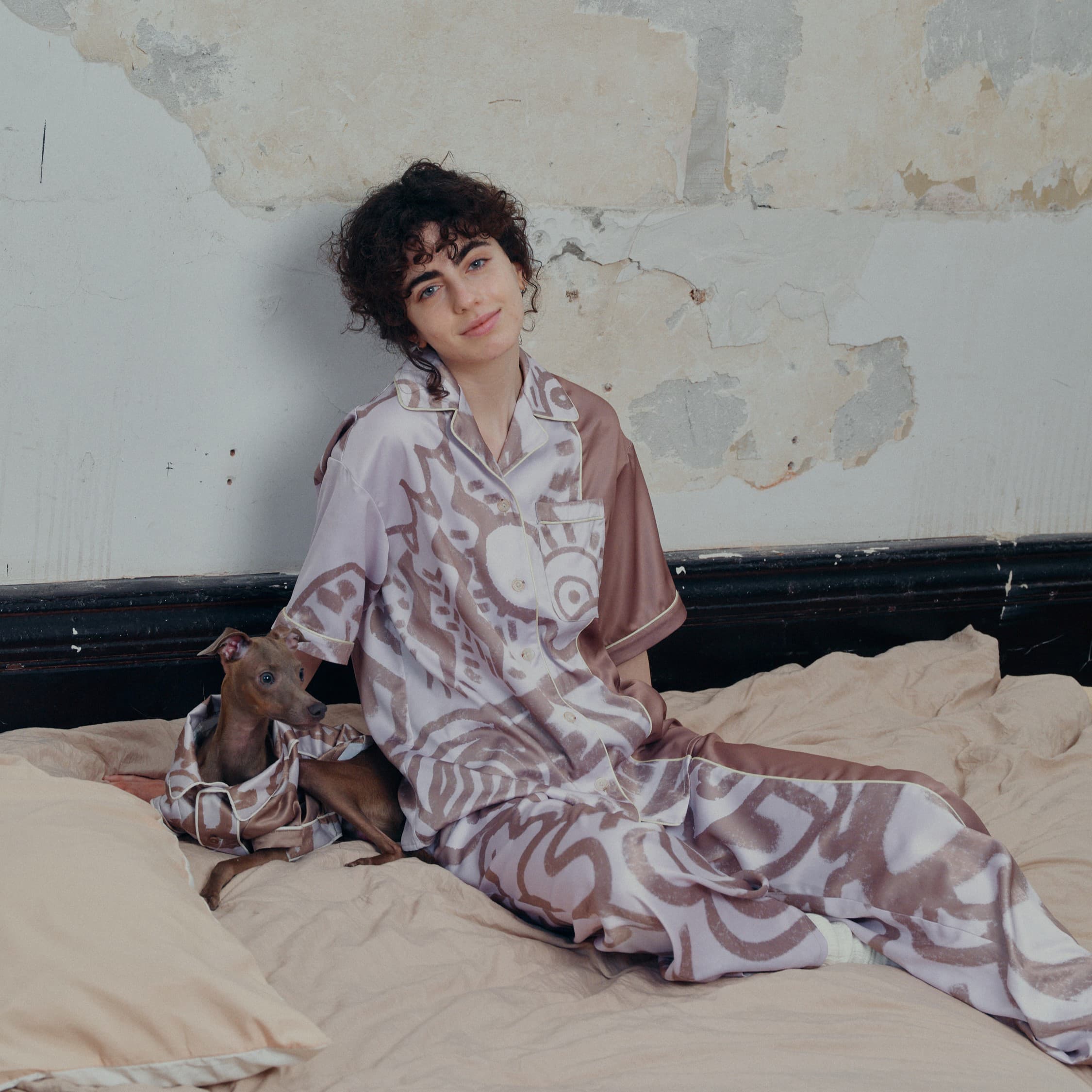 A smiling female model sits on a bed with a dog, both the owner and the dog wear short sleeve pajama sets.