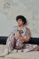 A male model sits on a bed with his dog, both the owner and the dog wear short sleeve pajama sets.