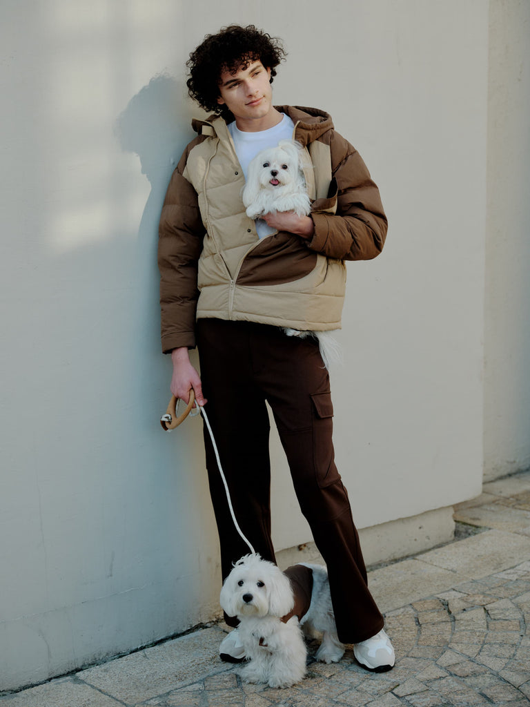 A male model is holding a dog that has been wearing a brown paded vest and holding a dog in his arms