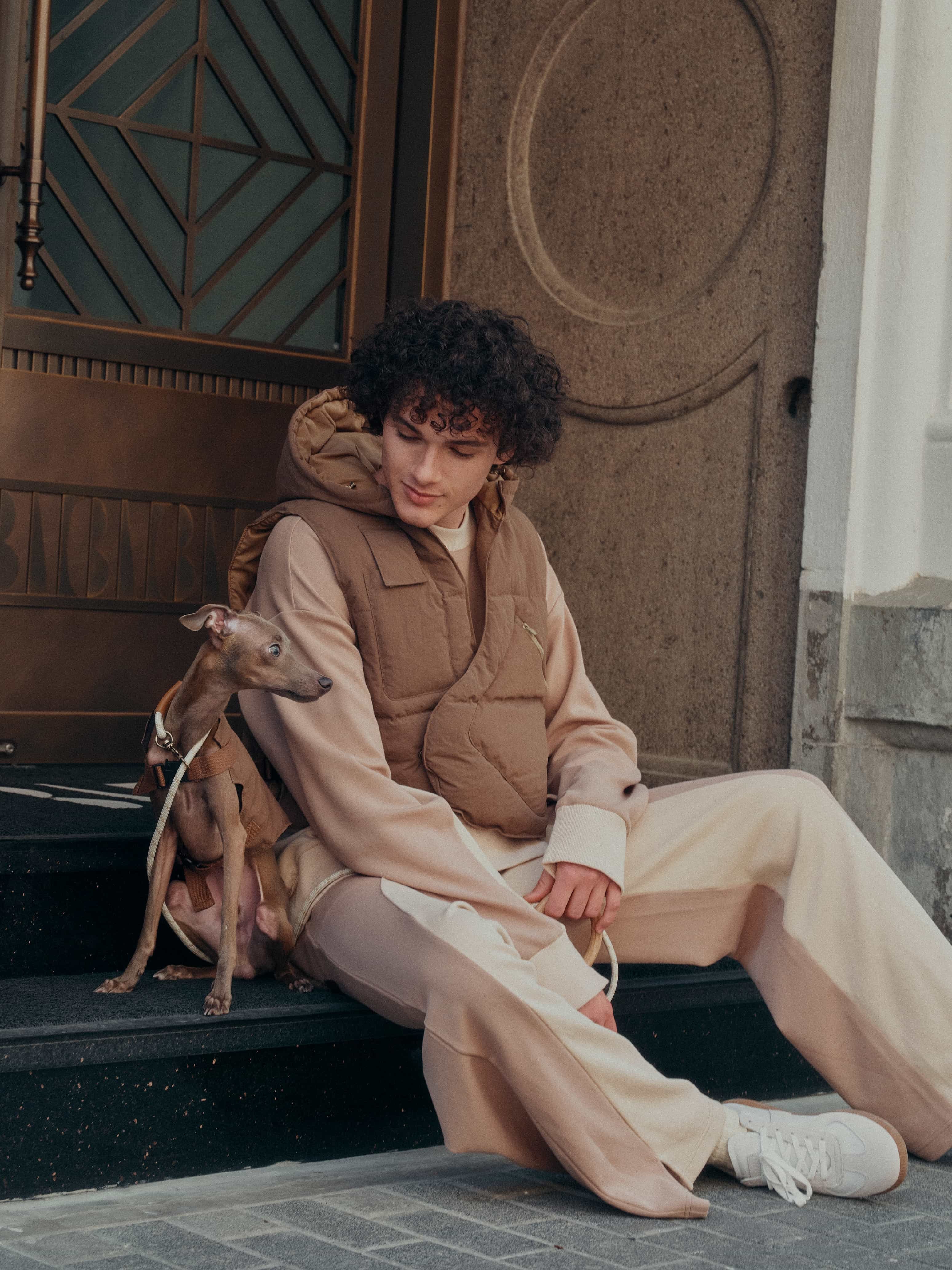 male model is sitting on the steps holding a dog on a leash. Both of them are wearing brown vests.