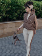 A female model, elegantly dressed in a brown vest and leggings, walks a dog on a leash.